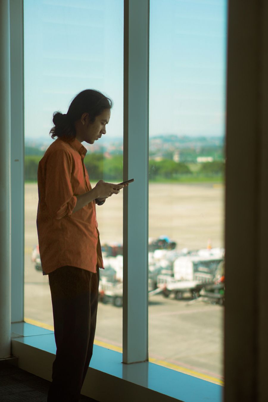 Man in orange shirt using a smartphone while standing at an airport window, overlooking the runway.