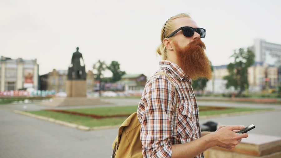 Hipster with a beard and sunglasses in a city square holding a smartphone. — O painel de partidas muda: portão achado, sala localizada, tudo no eSIM.